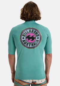 Rash guard verde azulado con mangas cortas que presenta un gran logo circular multicolor en la parte posterior, resaltando el nombre de la marca "Billabong Australia".