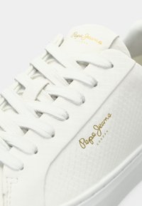 Hvite sneakers med teksturert materiale, flate lisser og en gull logo på siden. Slett overflate med et minimalistisk design.