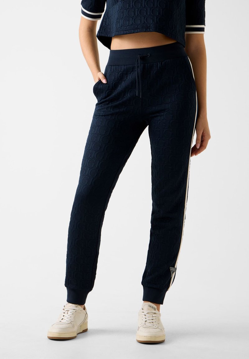 Frau in navyblauen, strukturierten Joggingshosen mit Seitenstreifen und weißen Turnschuhen, die mit einer Hand in der Tasche vor einem einfarbigen Hintergrund steht.