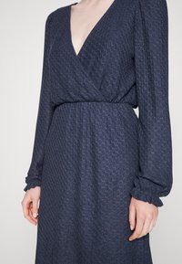 Vestido azul-marinho com design envelopado, tecido texturizado, mangas compridas com punhos franzidos e decote em V profundo; ajustado na cintura.