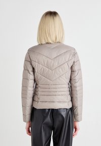 Lys beige pufferjakke med et chevron-mønster og horisontale sømmer på baksiden, med en tettsittende design og lange ermer.