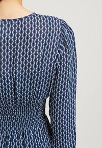 Vue arrière d'une personne portant une robe bleue à motifs avec des manches longues et une taille élastiquée froncée sur un fond uni.