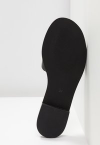 Semelle d'une chaussure noire avec talon et avant texturés, taille 37 visible, inclinée contre une surface blanche dans un décor minimaliste.