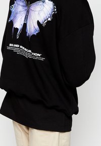 Sudadera XL negra con un gráfico de mariposa azul en la parte posterior, con texto blanco debajo. Textura de tela suave.