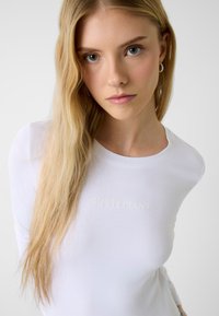 Bijela košulja s dugim rukavima i pripijenim krojem, s diskretnim, utisnutim natpisom "GUESS JEANS" na prsima. Mekana tekstura tkanine.