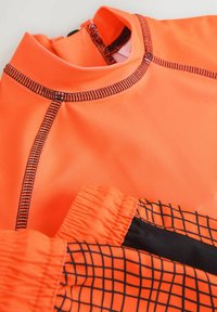Pomarańczowy rash guard wykonany z elastycznego materiału, z czarnymi akcentami i wzorem w kratkę, z wysokim kołnierzem i płaskimi szwami.