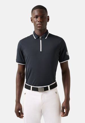 Man wearing black short-sleeve polo with white trim and zipper, white pants, and black belt with silver buckle standing against white background.