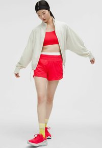 Sujetador deportivo rojo, pantalones cortos rojos a juego con cintura blanca, chaqueta ligera oversized, y zapatillas rosas con suelas grises. Calcetines a rayas amarillas.
