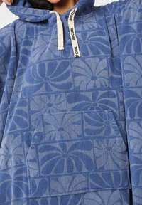 Poncho blu con cappuccio realizzato in morbido pile texturizzato con un motivo floreale. Presenta una tasca frontale e una chiusura con cordino.