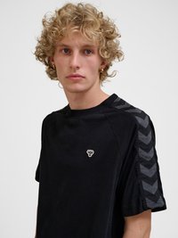 Noor inimene, kellel on lokkis blondid juuksed, kannab musta T-särki tumedate sarvkõrvade triipudega varrukatel ja väikesest valgest mesilase logost rinnal.