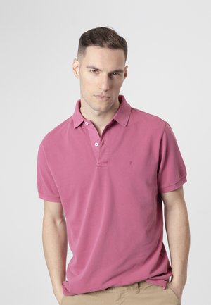 Hombre con cabello corto y oscuro, lleva una camisa polo rosa y pantalones beige, de pie con las manos en los bolsillos frente a un fondo liso.
