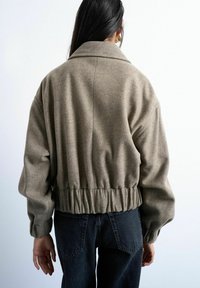 Personne aux cheveux longs et foncés porte une veste taupe courte avec une taille froncée et un jean foncé, vue de dos sur un fond uni.