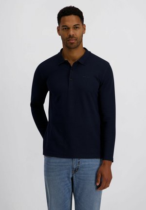 Mann steht vor einfachem Hintergrund, trägt ein marineblaues Langarm-Poloshirt und hellblaue Jeans, schaut nach vorne mit neutralem Gesichtsausdruck.