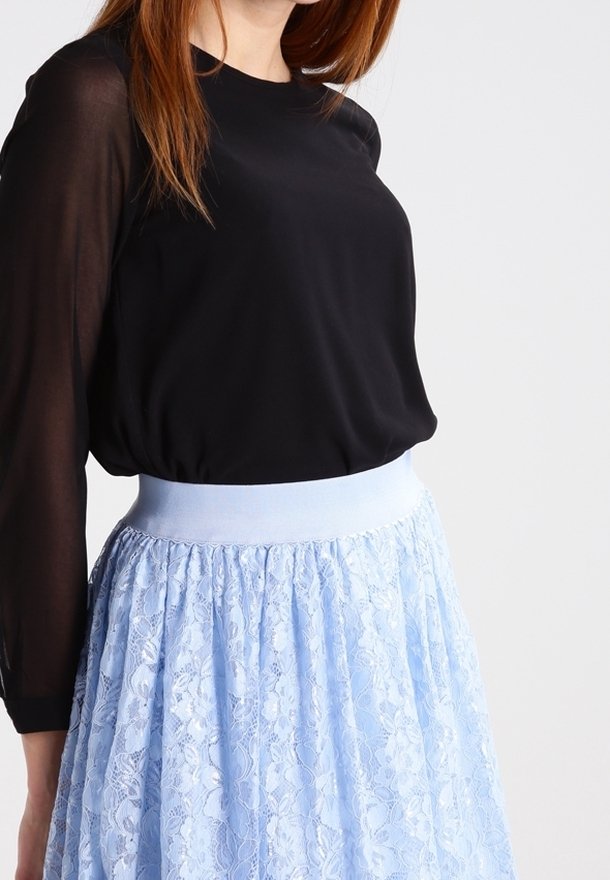 Donna che indossa una blusa nera a maniche lunghe e trasparente, infilata in una gonna di pizzo azzurro chiaro a vita alta, su uno sfondo bianco.