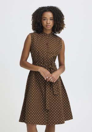 Vrouw met krullend haar, gekleed in een mouwloze bruine jurk met witte stippen, een knoopsluiting aan de voorkant en een ceintuur in de taille, staand tegen een effen achtergrond.