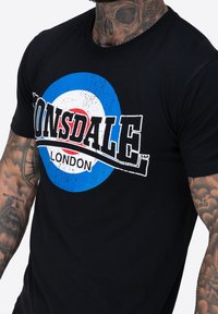 Must musta T-särk musta värviga, millel on suur Lonsdale London logo, sisaldades sinise ja valge sihikutega kujundust ning kulunud kirjete efekti. Lühikesed varrukad, tavaline lõige.