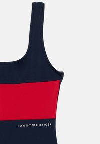 Μαγιό σε ναυτικό μπλε με κόκκινη οριζόντια ρίγα στο στήθος, με λείο ύφασμα και ορατές ραφές. Λογότυπο: "TOMMY HILFIGER" σε λευκό.