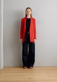 Blazer rosso sopra un top nero, abbinato a jeans scuri a vita alta. Scarpe rosse con una suola a contrasto. Design semplice e strutturato.