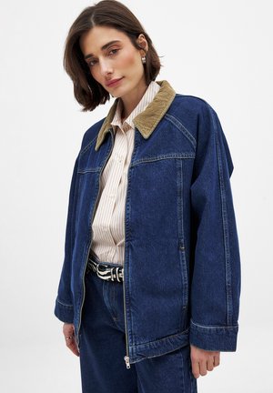 Denim jacket with a corduroy collar, featuring a zip closure, loose fit, and reinforced seams in dark blue. Worn over a striped shirt.
