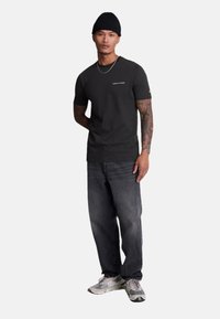 Tricou negru cu mânecă scurtă, cu textul "LYLE & SCOTT", asociat cu blugi închisi și adidași gri. Prezintă un guler cu nervuri și o pălărie beanie.