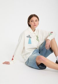 Jeune femme assise par terre portant une chemise blanche avec des motifs de cactus, champignons et fleurs, et un short en jean arrivant aux genoux.