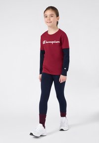 Burgunderfarget T-skjorte med korte ermer og hvit "Champion" logo, kombinert med marineblå leggings. Hvite sko og burgunder aksenter på sokkene. Jevne teksturer gjennomgående.