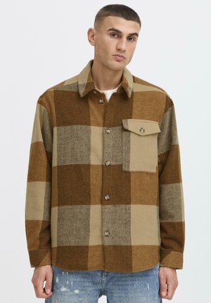 Uomo che indossa una camicia a quadri grandi marrone e beige con taschino sul petto, abbinata a jeans azzurri strappati, in piedi contro uno sfondo bianco.