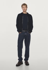 Dunkelgrau zipfeliger Hoodie über einem marineblauen Shirt, kombiniert mit blauen Jeans und braunen Stiefeln. Schlichtes Design mit lockerem Schnitt und ohne sichtbare Muster.