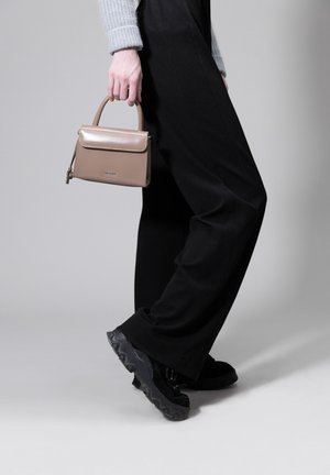 Tan leather handbag with a rectangular shape and a front flap, held in a hand. Black wide-leg trousers and chunky black shoes are also visible.