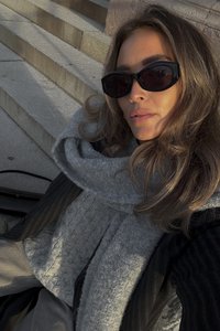 Grauer strukturierter Pullover, überzogen mit einem schwarzen Mantel und einem dicken grauen Schal. Die Person trägt große schwarze Sonnenbrillen und hat hellbraunes Haar.