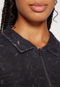 Chaqueta de mezclilla negra con cuello y cierre de cremallera, que presenta una superficie texturizada con pequeñas motas y un diseño ajustado en general.