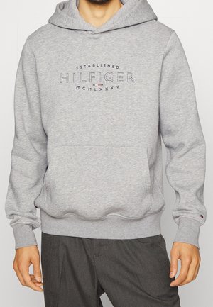 Mężczyzna ubrany w jasnoszarą bluzę z kapturem Tommy Hilfiger z przednią kieszenią i napisem logo "Established Hilfiger MCMLXXXV" na piersi.