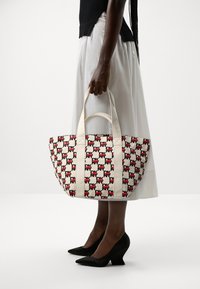 Sac en toile présentant un motif géométrique rouge et noir, des poignées crème, une forme arrondie et des détails de coutures prononcés.