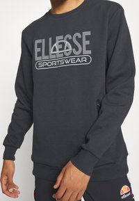 Sweat-shirt gris foncé avec col rond, manches longues et logo à l'avant portant le texte "SPORTSWEAR". Comprend un détail de poche latérale.