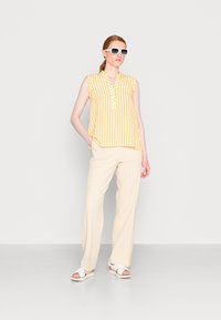 Blusa sem mangas às riscas amarelas e brancas com colarinho, combinada com calças largas em creme e sandálias brancas. Modelo usa óculos de sol oversized brancos.