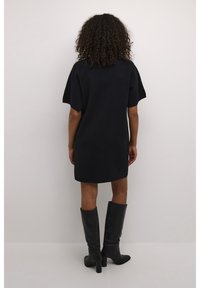 Femme aux cheveux bouclés debout, portant une robe noire courte et des bottes noires à talons montant jusqu'aux genoux, face à un mur blanc uni.