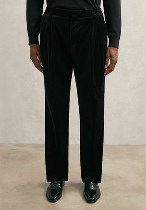 Musta sametist püksid, millel on mugav lõige, voldid, elastne vöökoht ja läikivad mustad kingad. Sile tekstuur ja ühtlane värv.