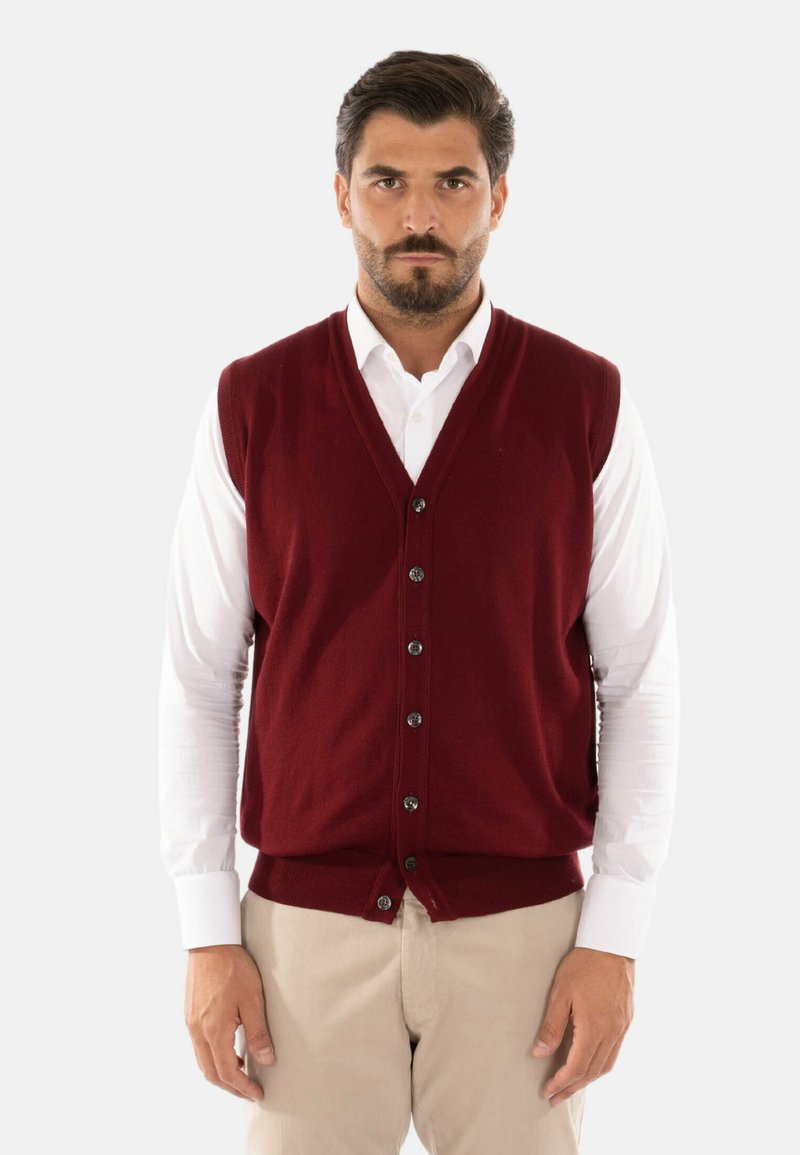 Gilet a maglia bordeaux con scollo a V, cinque bottoni neri e orlo a coste, indossato sopra una camicia bianca a maniche lunghe e pantaloni beige.