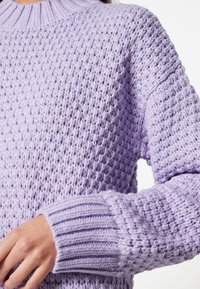 Pull en tricot violet clair avec un motif texturé, des poignets côtelés et un col rond haut. La laine douce et épaisse ajoute de la dimension.