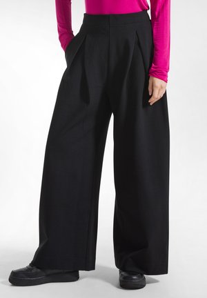 Persoon draagt zwarte wijdvallende broek, zwarte schoenen en een felroze top met lange mouwen, met één hand in de zak en de andere ontspannen.