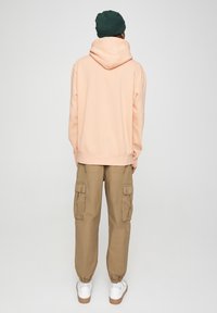 Felpa color pesca chiaro con cappuccio regolabile, abbinata a pantaloni cargo color khaki con tasche laterali e polsini elastici, completata da sneakers bianche.