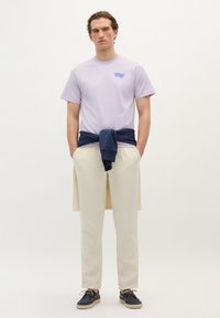 Lyslilla kortærmet t-shirt med et blåt grafik, beige bukser, marineblå sweater bundet omkring livet, og sorte sko med snørebånd.