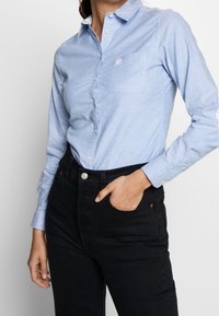 Camicia azzurra chiara con colletto, caratterizzata da una singola tasca sul petto, chiusura a bottoni e maniche lunghe. Abbinata a jeans neri a vita alta.