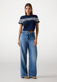 T-shirt blu navy cropped con strisce bianche e design grafico, abbinato a jeans a gamba larga di un blu chiaro. Tessuto denim liscio, vestibilità rilassata.