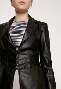 Černá kožená blazer s přiléhavým pasem, klopovými reverzy, zipem a hladkým povrchem, spárovaná s pruhovaným topem pod tím.