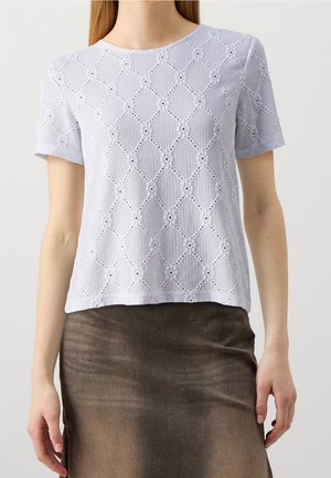 Mujer con una blusa blanca de encaje de manga corta con un patrón de diamantes y una falda marrón de cintura alta.