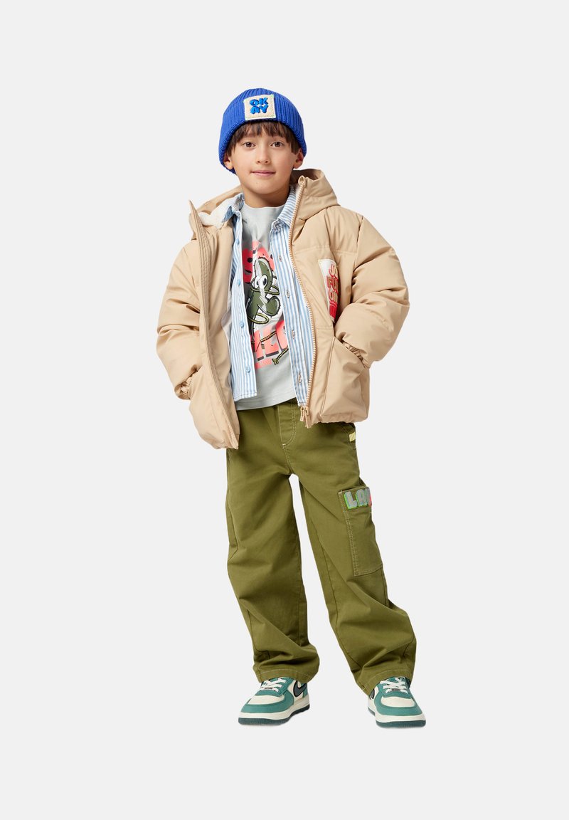 Beige pufferjack, lichtblauwe gestreepte shirt, graphic t-shirt, olijfgroene cargobroek met een patch, blauwe gebreide beanie, groene sneakers.
