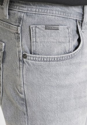 Sive traperice s diskretnom teksturom, koje imaju stražnji džep i malu crnu oznaku. Standardni kroj s kontrastnim šavovima.