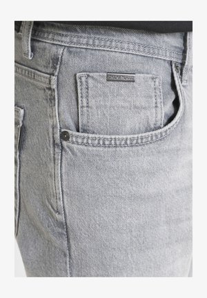 Sive traperice s diskretnom teksturom, koje imaju stražnji džep i malu crnu oznaku. Standardni kroj s kontrastnim šavovima.