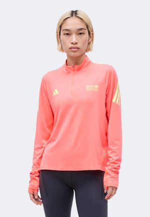 Persoon draagt een koraalrode sporttop met lange mouwen met BMW Berlin Marathon- en Adidas-logo's, gecombineerd met zwarte leggings, staand tegen een witte achtergrond.